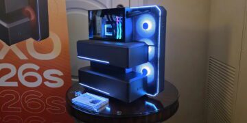 نمای سه‌رخ از کیس کامپیوتر پیشرفته با ساختار چندبخشی پله‌ای، پنل جلویی مشبک با نورپردازی LED آبی، سه فن خنک‌کننده دایره‌ای، پنل شیشه‌ای کناری و نمایش ماژول سخت‌افزاری داخلی در محیط نمایشگاهی.