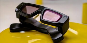 نمای استودیویی عینک هوشمند Snapchat Spectacles با طراحی ضخیم و صنعتی، لنزهای رنگی مایل به بنفش، فریم مشکی مات و ماژول‌های دوربین یکپارچه در گوشه‌های فریم؛ محصول روی پایه‌ی نگهدارنده‌ی مشکی و سطح زرد براق قرار گرفته است.