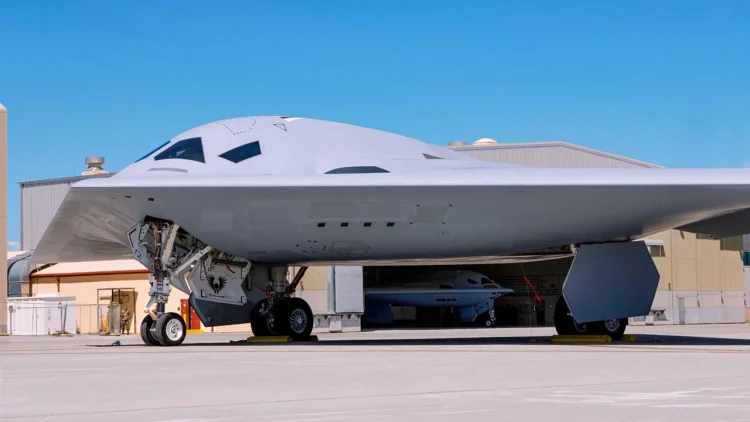 نمای روبه‌روی بمب‌افکن رادارگریز B-21 Raider در داخل آشیانه با پرچم بزرگ ایالات متحده در پس‌زمینه