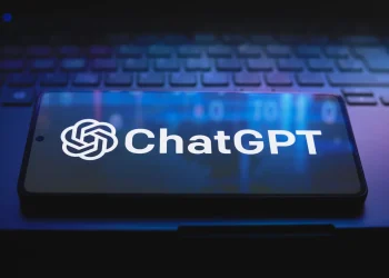 نمایش لوگو و نام ChatGPT بر روی صفحه نمایش یک گوشی هوشمند که روی کیبورد یک لپ‌تاپ قرار دارد؛ تصویری نمادین از دسترسی گسترده به هوش مصنوعی مولد.