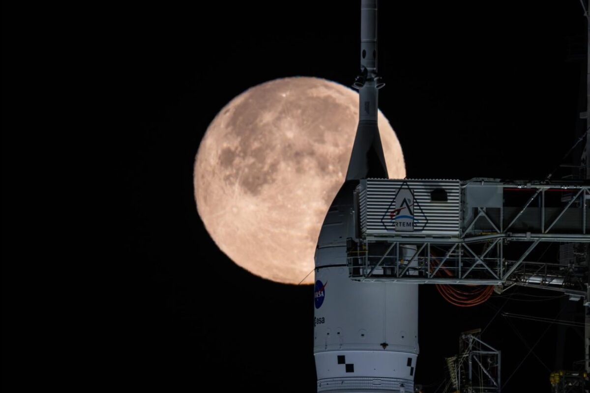 نمایی نزدیک از موشک و سازه پرتاب ناسا روی سکوی پرتاب، با ماه کامل در پس‌زمینه آسمان شب؛ ترکیبی سینمایی که هم‌زمان فناوری فضایی و پدیده نجومی را نمایش می‌دهد.