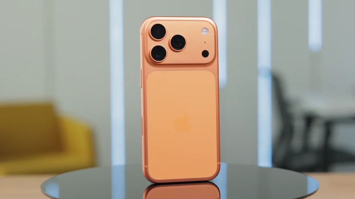 نمای پشت یک گوشی iPhone با طراحی مدرن و بدنه فلزی در رنگ روشن، مجهز به ماژول دوربین سهگانه مربعیشکل با لنزهای بزرگ، که روی سطحی براق قرار گرفته و پسزمینهای محو از فضای داخلی مدرن دارد؛ تصویری که به تغییرات سختافزاری احتمالی آیفونهای آینده و تصمیمات اپل در زنجیره تأمین اشاره دارد.