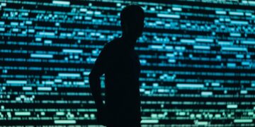 سایه تیره یک انسان در مقابل صفحه‌ای پر از کدهای دیجیتالی آبی‌رنگ، نمادی از کاربران ناشناس یا هکرها.