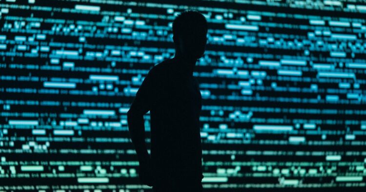 سایه تیره یک انسان در مقابل صفحه‌ای پر از کدهای دیجیتالی آبی‌رنگ، نمادی از کاربران ناشناس یا هکرها.