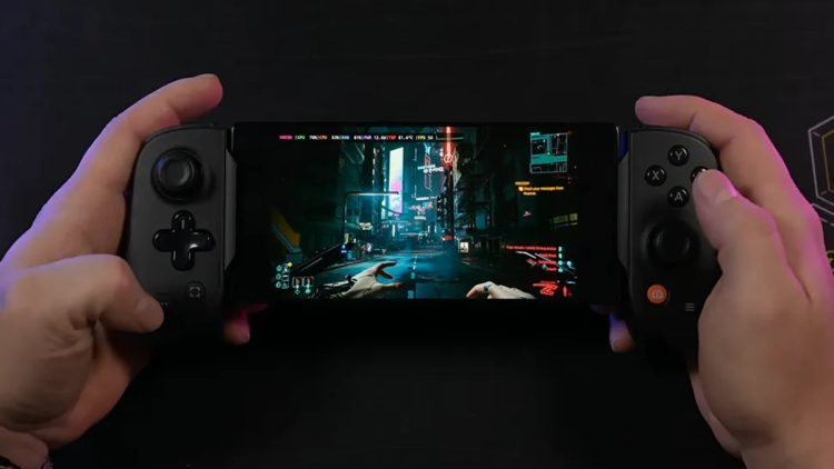 نمای یک گوشی هوشمند که به کنترلر گیمینگ فیزیکی دوطرفه متصل شده و بازی Cyberpunk 2077 را در حالت نمای اول‌شخص اجرا می‌کند. تصویر خیابان‌های نئونی شهر Night City با نورپردازی سایبرپانکی روی نمایشگر دیده می‌شود و این صحنه اجرای بازی AAA روی موبایل را نمایش می‌دهد.
