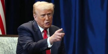 ترامپ با کت‌وشلوار تیره و کراوات قرمز در حال اشاره با دست، در فضای داخلی رسمی با پرده تیره و پرچم در پس‌زمینه