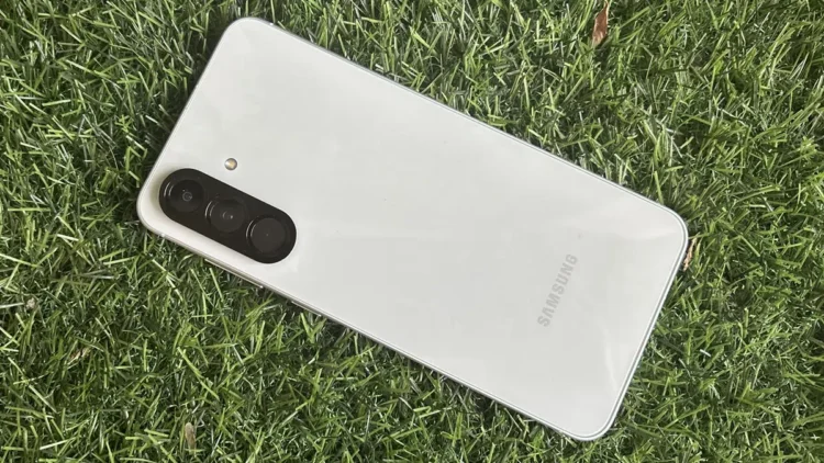 نمای واقعی از پشت گوشی Samsung Galaxy A56 با بدنه روشن، ماژول دوربین سه‌گانه عمودی و فریم تخت که روی سطح چمن قرار گرفته و جزئیات طراحی فیزیکی دستگاه را نمایش می‌دهد.
