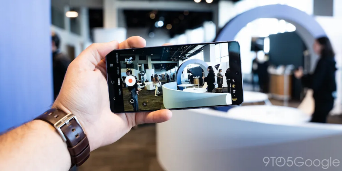 فردی که یک گوشی هوشمند را در دست گرفته و در حال فیلمبرداری از یک محیط نمایشگاهی است؛ رابط کاربری دوربین روی صفحه نمایش دیده میشود.