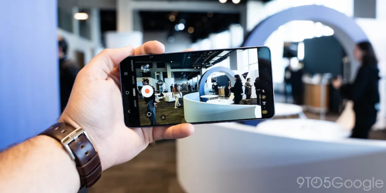 فردی که یک گوشی هوشمند را در دست گرفته و در حال فیلم‌برداری از یک محیط نمایشگاهی است؛ رابط کاربری دوربین روی صفحه نمایش دیده می‌شود.