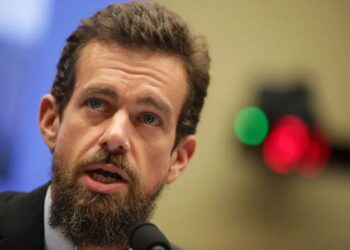 نمای نزدیک از جک دورسی پشت میکروفون در یک رویداد رسمی با کت‌وشلوار تیره؛ پس‌زمینه محو با نورهای دایره‌ای سبز و قرمز که فضای کنفرانس/نشست خبری را القا می‌کند