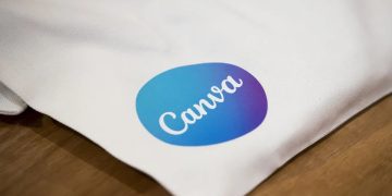 نمای نزدیک از لوگوی Canva چاپ‌شده روی سطح پارچه‌ای سفید که روی میز چوبی قرار گرفته است. بافت پارچه و چاپ رنگی لوگو به‌وضوح دیده می‌شود و تصویر به استفاده فیزیکی از برند Canva در محصولات تبلیغاتی اشاره دارد.