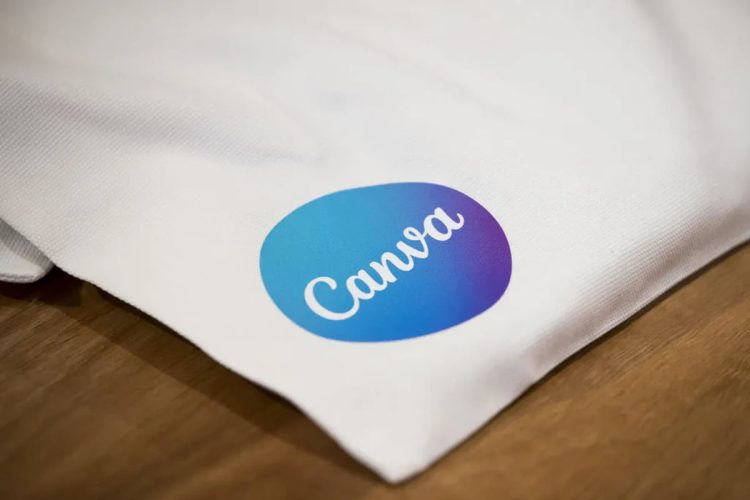 نمای نزدیک از لوگوی Canva چاپشده روی سطح پارچهای سفید که روی میز چوبی قرار گرفته است. بافت پارچه و چاپ رنگی لوگو بهوضوح دیده میشود و تصویر به استفاده فیزیکی از برند Canva در محصولات تبلیغاتی اشاره دارد.