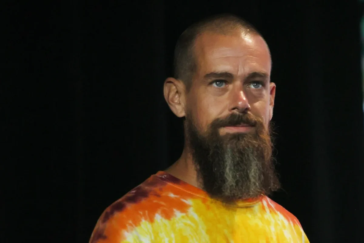 پرتره نیم‌تنه از جک دورسی با تی‌شرت تای‌دای نارنجی و زرد در پس‌زمینه مشکی؛ نورپردازی استودیویی و کنتراست بالا با تمرکز بر بافت لباس و ریش بلند