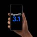 دستی که یک گوشی هوشمند را با لوگوی HyperOS 3.1 در پس‌زمینه مشکی نگه داشته است.