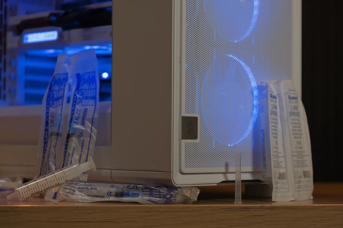 نمای نزدیک از قسمت پایینی کیس کامپیوتر و سرنگ‌های پزشکی قرار گرفته روی لبه میز.