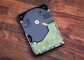 نمای بالایی از یک هارد دیسک ۳.۵ اینچی (HDD) با قاب فلزی بازنشده که روی سطح چوبی قرار گرفته است؛ برد مدار چاپی (PCB) سبز، کانکتورهای SATA و بخش مکانیکی داخلی هارد قابل مشاهده هستند.