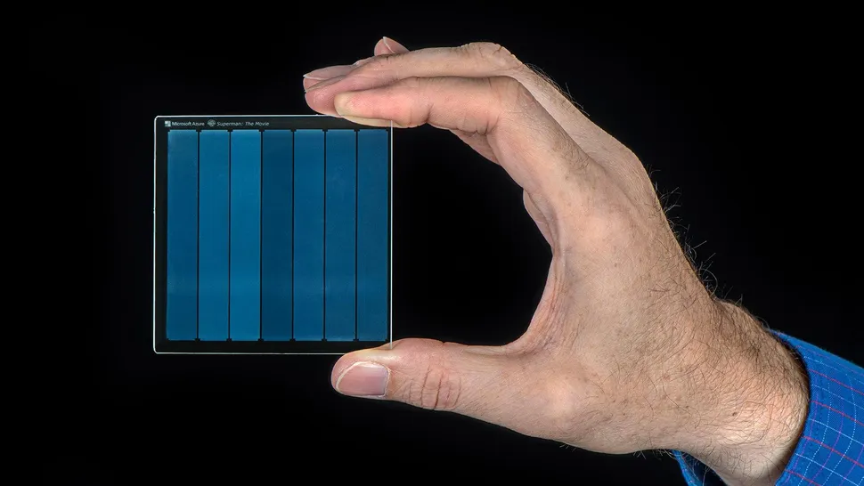 دستی که یک لوح شیشه‌ای شفاف و کوچک (تکنولوژی ذخیره‌سازی داده) را زیر نور مستقیم یک چراغ قوه برای بررسی نگه داشته است.