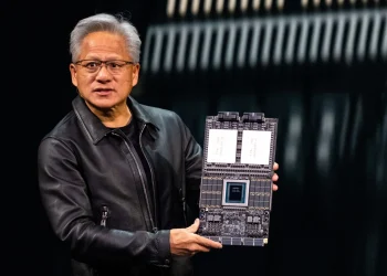 نمای نزدیک از جن‌سون هوانگ که یک برد مدار چاپی بزرگ و پیچیده (تراشه شتاب‌دهنده) را با دو دست برای نمایش نگه داشته است.