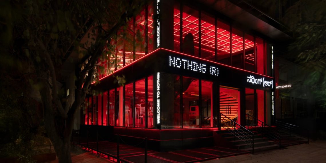 نمای بیرونی فروشگاه رسمی Nothing ® در هند، با معماری مدرن شیشه‌ای، نورپردازی قرمز چشمگیر در فضای داخلی، و لوگوی LED برند Nothing روی نمای ساختمان. طراحی مینیمال فروشگاه با ورودی پلکانی، المان‌های صنعتی و فضای شهری اطراف، هویت بصری متمایز برند را برجسته می‌کند.