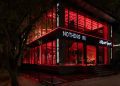 نمای بیرونی فروشگاه رسمی Nothing ® در هند، با معماری مدرن شیشه‌ای، نورپردازی قرمز چشمگیر در فضای داخلی، و لوگوی LED برند Nothing روی نمای ساختمان. طراحی مینیمال فروشگاه با ورودی پلکانی، المان‌های صنعتی و فضای شهری اطراف، هویت بصری متمایز برند را برجسته می‌کند.
