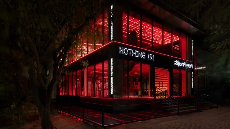 نمای بیرونی فروشگاه رسمی Nothing ® در هند، با معماری مدرن شیشه‌ای، نورپردازی قرمز چشمگیر در فضای داخلی، و لوگوی LED برند Nothing روی نمای ساختمان. طراحی مینیمال فروشگاه با ورودی پلکانی، المان‌های صنعتی و فضای شهری اطراف، هویت بصری متمایز برند را برجسته می‌کند.