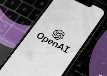 لوگوی OpenAI روی صفحه‌نمایش یک گوشی هوشمند که بر روی کیبورد لپ‌تاپ قرار دارد.