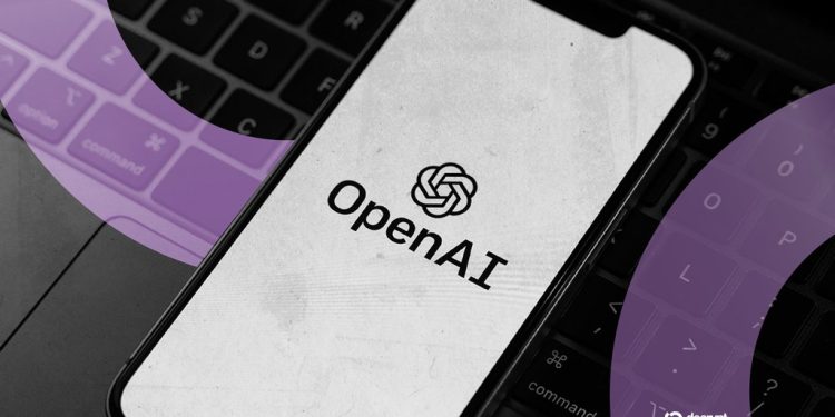 لوگوی OpenAI روی صفحهنمایش یک گوشی هوشمند که بر روی کیبورد لپتاپ قرار دارد.