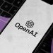 لوگوی OpenAI روی صفحه‌نمایش یک گوشی هوشمند که بر روی کیبورد لپ‌تاپ قرار دارد.