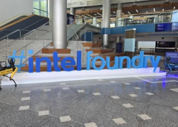 نمای داخلی یک ساختمان مدرن با لوگوی بزرگ Intel Foundry به رنگ آبی در مرکز سالن؛ دو ربات چهارپا در دو طرف نوشته قرار دارند و راه‌پله‌ها و نمایشگرهای دیجیتال در پس‌زمینه دیده می‌شود