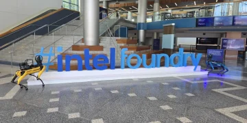 نمای داخلی یک ساختمان مدرن با لوگوی بزرگ Intel Foundry به رنگ آبی در مرکز سالن؛ دو ربات چهارپا در دو طرف نوشته قرار دارند و راهپلهها و نمایشگرهای دیجیتال در پسزمینه دیده میشود