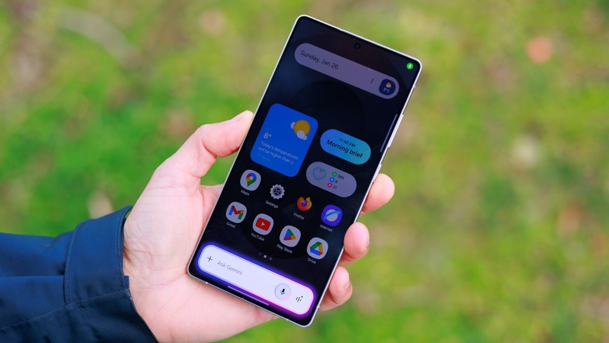 نمای جلوی گوشی سامسونگ در دست با ویجت‌های آب‌وهوا و آیکون برنامه‌ها در محیط باز.