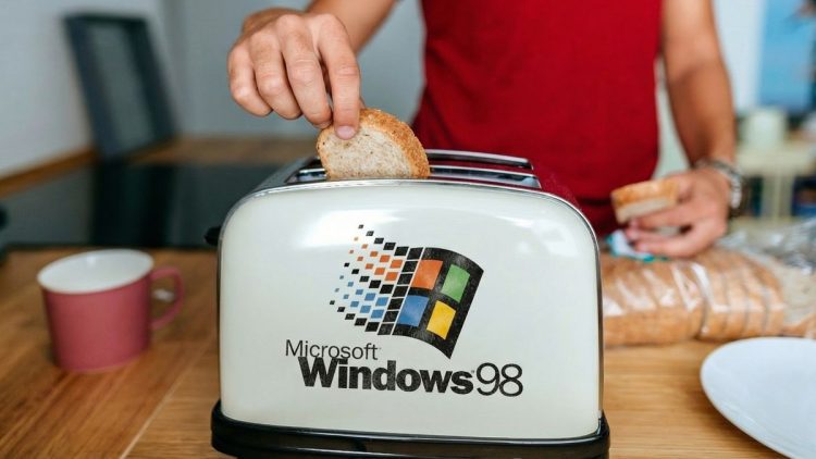 دست فردی در حال قرار دادن نان در توستری که لوگوی بزرگ Microsoft Windows 98 روی بدنه آن نقش بسته است.