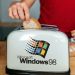 دست فردی در حال قرار دادن نان در توستری که لوگوی بزرگ Microsoft Windows 98 روی بدنه آن نقش بسته است.