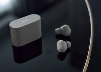 هدفون بی‌سیم Sony XM6 به رنگ نقره‌ای در کنار کیس شارژ روی میز براق.