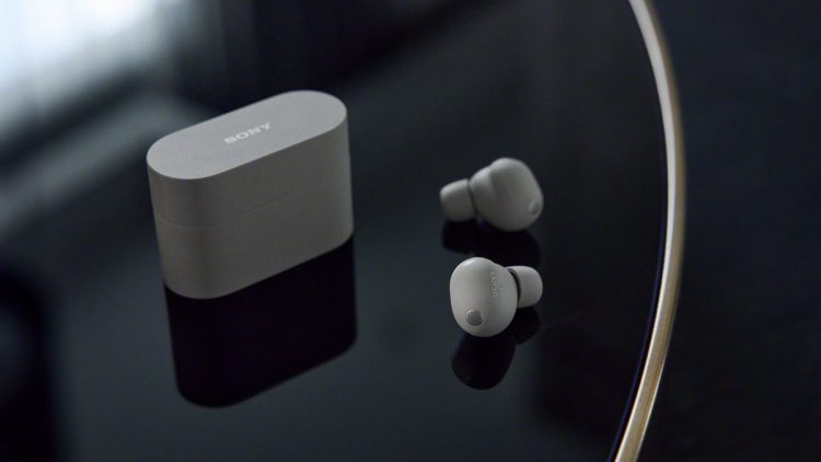 هدفون بی‌سیم Sony XM6 به رنگ نقره‌ای در کنار کیس شارژ روی میز براق.