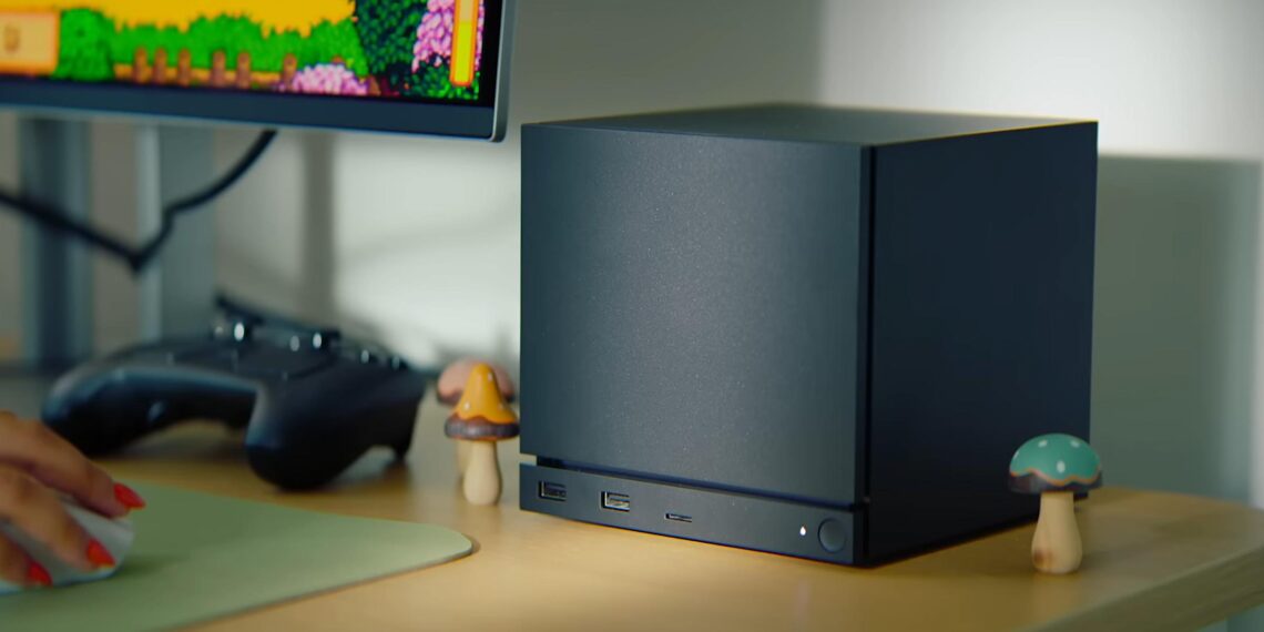 کنسول Steam Machine با طراحی مکعبی مشکی روی میز، مجهز به نوار LED آبی در پنل جلویی، در کنار نمایشگر متصل که محیط کاربری Steam را نمایش می‌دهد.