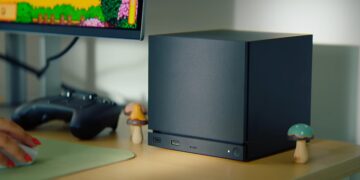 کنسول Steam Machine با طراحی مکعبی مشکی روی میز، مجهز به نوار LED آبی در پنل جلویی، در کنار نمایشگر متصل که محیط کاربری Steam را نمایش می‌دهد.