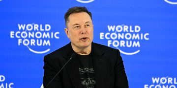 نمای یک سخنرانی رسمی در رویداد World Economic Forum با پس‌زمینه آبی و لوگوی تکرارشونده این رویداد جهانی. یک سخنران با پوشش رسمی در پشت تریبون و میکروفن قرار دارد و فضای تصویر، حال‌وهوای کنفرانس اقتصادی بین‌المللی، سیاست‌گذاری و فناوری را منتقل می‌کند.