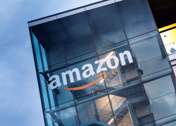 نمای بیرونی یک ساختمان اداری مدرن با نمای شیشه‌ای که لوگوی بزرگ Amazon به‌همراه فلش نارنجی معروف این برند روی شیشه نصب شده است؛ سازه داخلی شامل راه‌پله‌ها و نورپردازی صنعتی دیده می‌شود و تصویر بر هویت شرکتی و معماری دفتر آمازون تمرکز دارد.