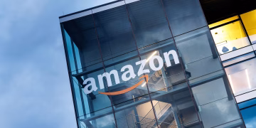 نمای بیرونی یک ساختمان اداری مدرن با نمای شیشه‌ای که لوگوی بزرگ Amazon به‌همراه فلش نارنجی معروف این برند روی شیشه نصب شده است؛ سازه داخلی شامل راه‌پله‌ها و نورپردازی صنعتی دیده می‌شود و تصویر بر هویت شرکتی و معماری دفتر آمازون تمرکز دارد.