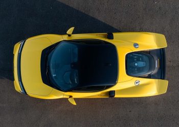 نمای بالا از یک خودروی اسپرت زرد Ferrari با موتور میانی که روی سطح آسفالت قرار دارد و محفظه موتور از پشت شیشه شفاف دیده می‌شود.