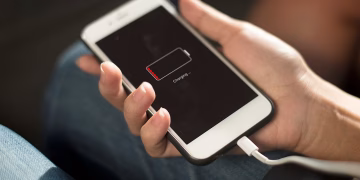 نمای نزدیک از یک گوشی آیفون متصل به کابل شارژ که روی صفحه آن آیکن باتری با سطح شارژ بسیار پایین و وضعیت Charging نمایش داده شده و به مشکل خالی‌شدن سریع باتری اشاره دارد.