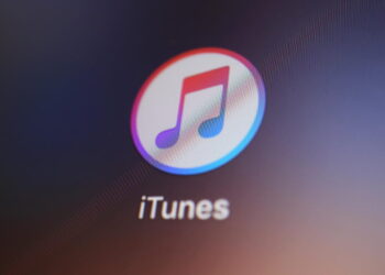 نمای نزدیک از آیکن اپلیکیشن iTunes با نماد نت موسیقی رنگی داخل یک دایره سفید، که روی صفحه‌نمایش دستگاه دیجیتال با پس‌زمینه تیره نمایش داده شده است.