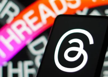نمای نزدیک از لوگوی اپلیکیشن Threads (تردز) روی نمایشگر گوشی هوشمند در پیش‌زمینه، با پس‌زمینه تار و رنگی شامل نوشته THREADS به رنگ نارنجی و بنفش — نمایش شاخصی از هویت بصری اپلیکیشن شبکه اجتماعی Threads