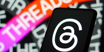 نمای نزدیک از لوگوی اپلیکیشن Threads (تردز) روی نمایشگر گوشی هوشمند در پیش‌زمینه، با پس‌زمینه تار و رنگی شامل نوشته THREADS به رنگ نارنجی و بنفش — نمایش شاخصی از هویت بصری اپلیکیشن شبکه اجتماعی Threads