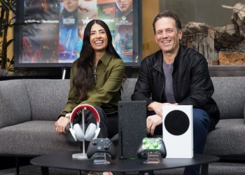 فیل اسپنسر و آشا شارما نشسته روی مبل در فضای معرفی محصولات گیمینگ، با نمایش کنسول Xbox، کنترلرها و هدست، در پس‌زمینه‌ای مرتبط با بازی‌های ویدیویی و اکوسیستم Xbox