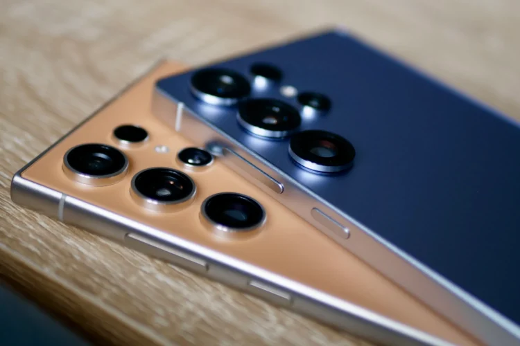 نمای نزدیک از پشت دو گوشی Samsung Galaxy S24 Ultra و Galaxy S25 Ultra که روی سطح چوبی قرار گرفتهاند؛ رنگ نارنجی/برنزی و آبی تیره بدنه در کنار ماژول دوربین چندگانه، تفاوت طراحی و چینش لنزها را بهوضوح نشان میدهد.