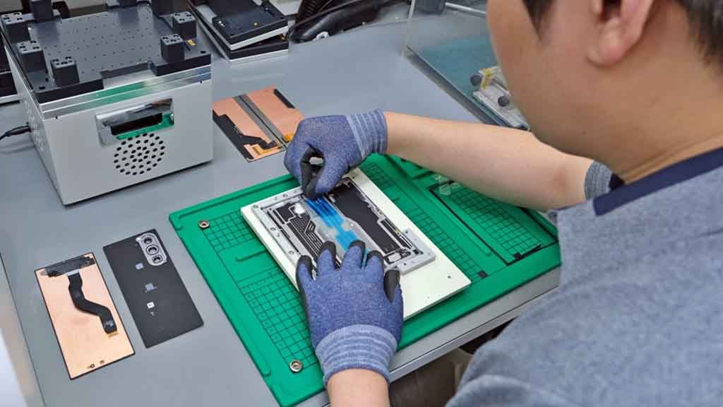 تکنسینی با دستکش‌های محافظ در حال تعمیر قطعات داخلی یک دستگاه هوشمند سامسونگ روی میز کار.