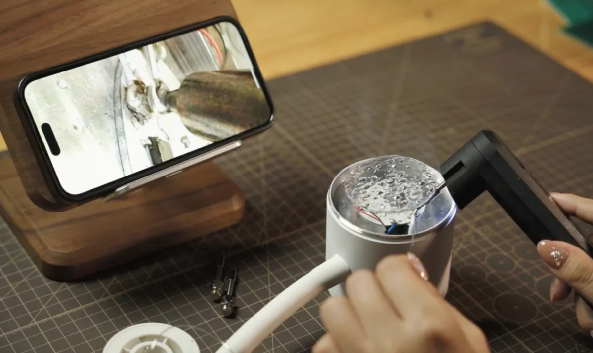 کاربر در حال مونتاژ و لحیمکاری یک ربات کوچک آموزشی با هویهی دوربیندار، همزمان با مشاهدهی جزئیات کار روی صفحهی گوشی