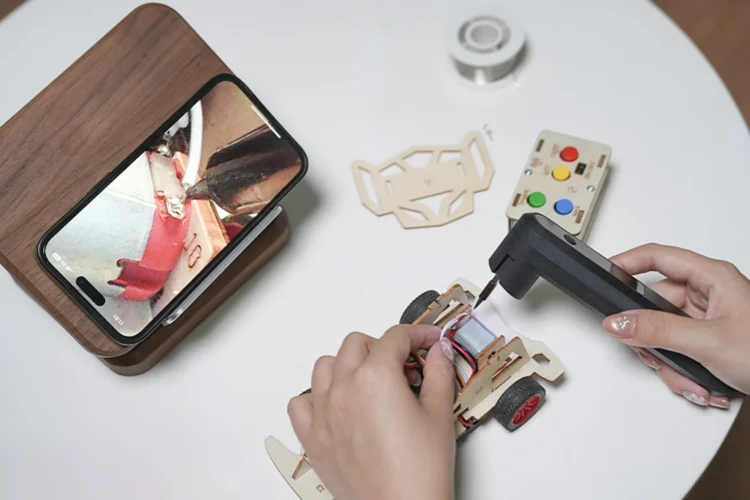 نمای نزدیک از لحیمکاری یک قطعه الکترونیکی که تصویر آن بهصورت زنده روی صفحهی تلفن هوشمند کنار میز کار نمایش داده میشود.